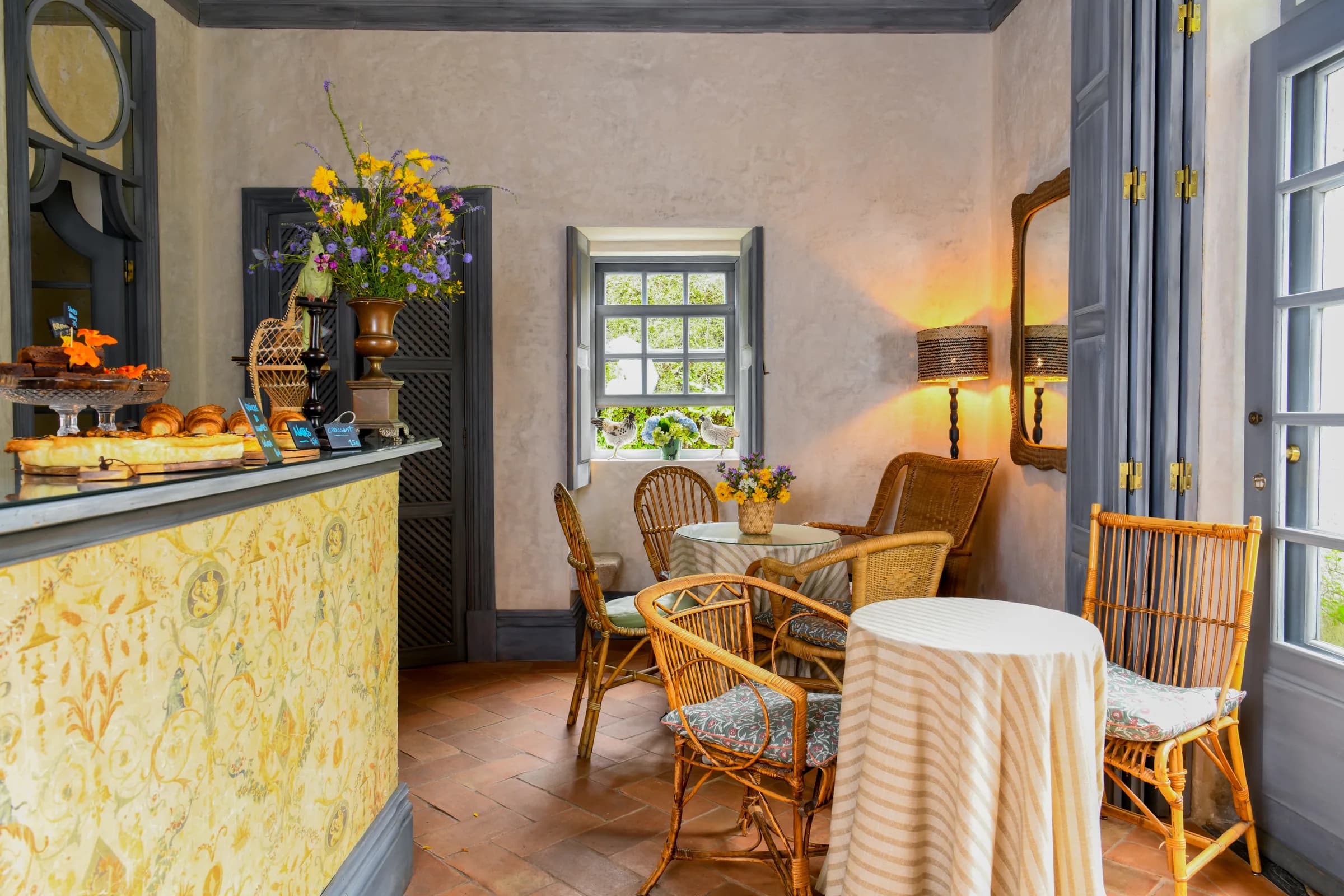 O balcão do café vestido com flores silvestres e pastelaria, cadeiras de verga junto à janela aberta