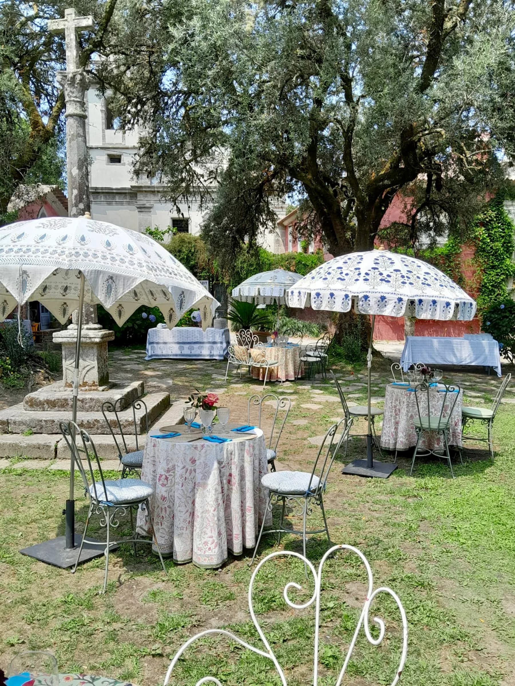 Café de Cabanas — guarda-sóis estampados sobre mesas postas no jardim, junto à antiga cruz e à torre da igreja